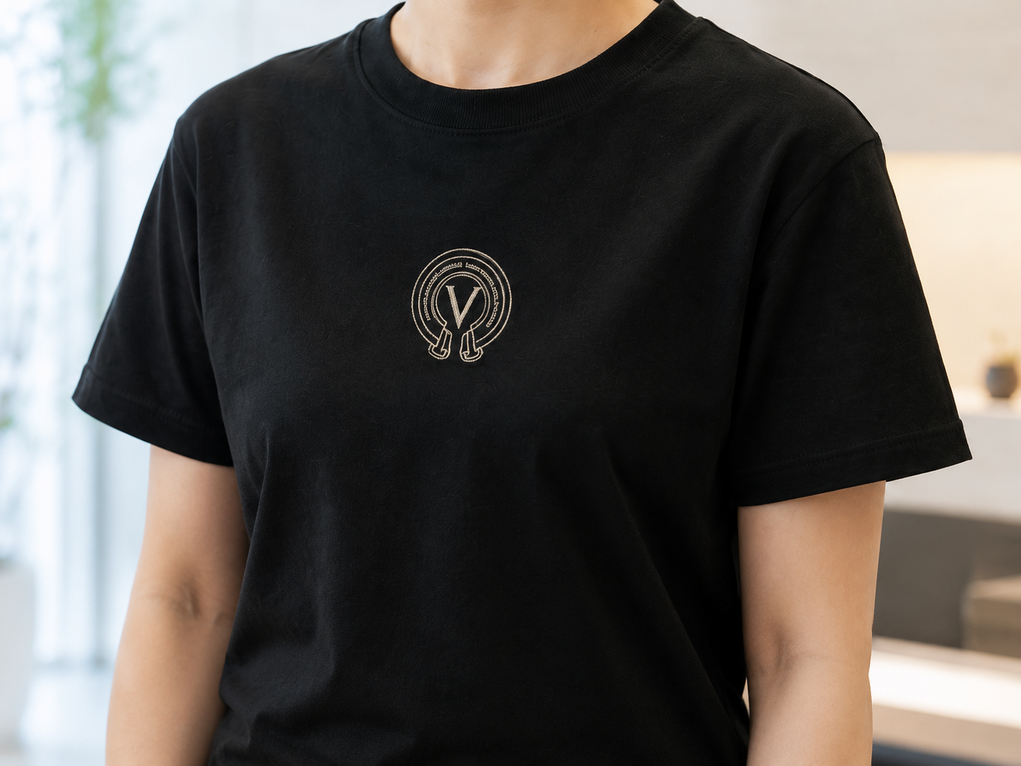 黒のTシャツの胸元に、生成りの糸で細く刺した V のモノグラム